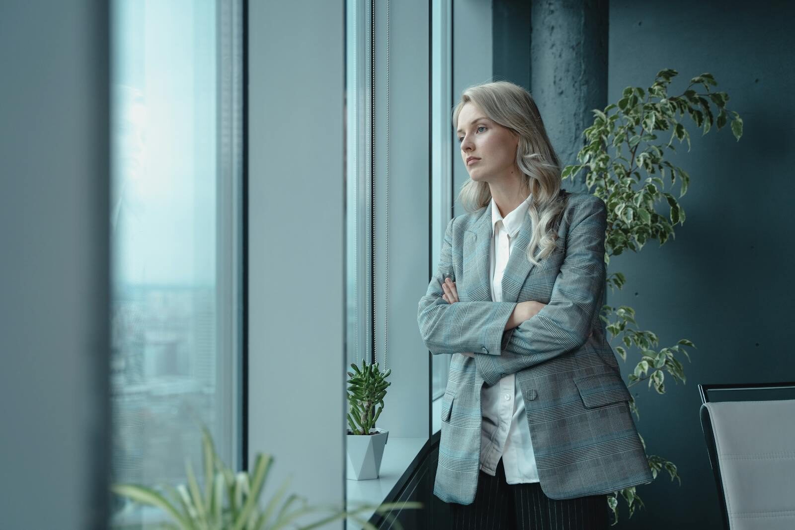 Professional businesswoman in elegant attire looks thoughtfully out a large office window.
